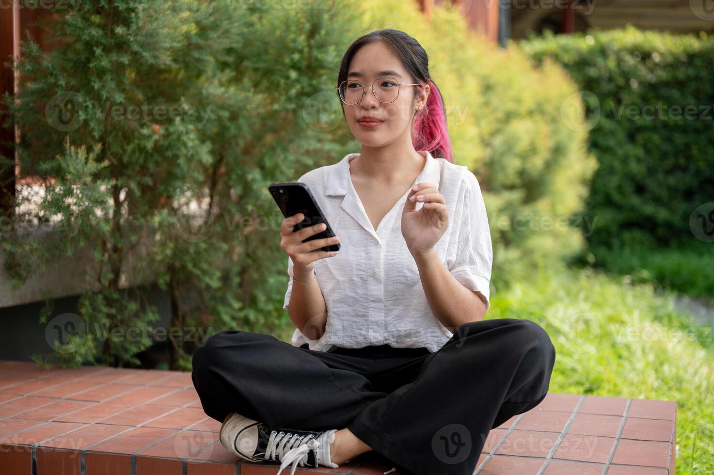 A young Asian woman in casual wear and glasses sits on a brick bench in a park using her smartphone. photo
