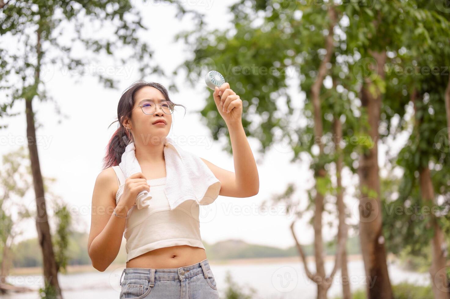 A woman holds a small fan while standing outdoors in a park, cooling herself down on a hot day. photo