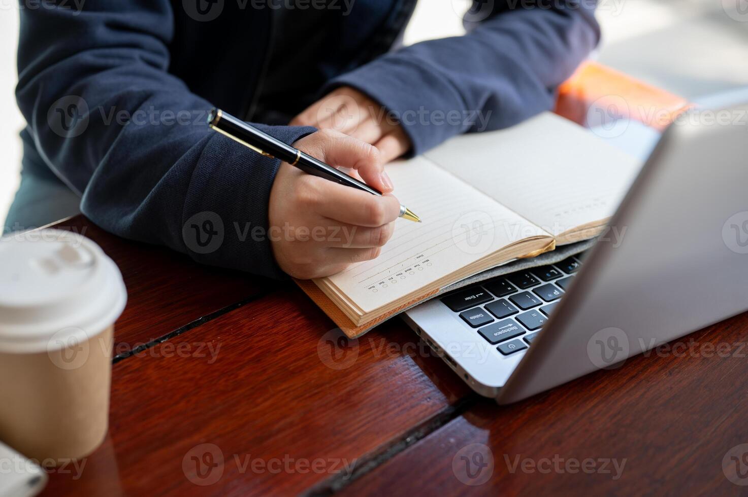 A woman is jotting down something in a notebook while working on her laptop at a table in a cafe. photo