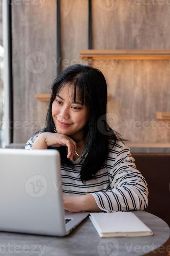A happy Asian woman is working remotely at a cafe co-working space, looking at her laptop screen. photo