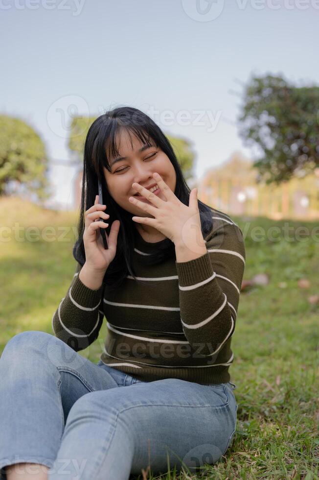 A cheerful young Asian woman enjoying talking on the phone with her friend while relaxing in a park. photo