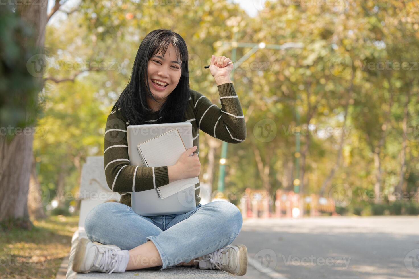A cheerful Asian female student is sitting on bench in a green park with her laptop and notebook. photo