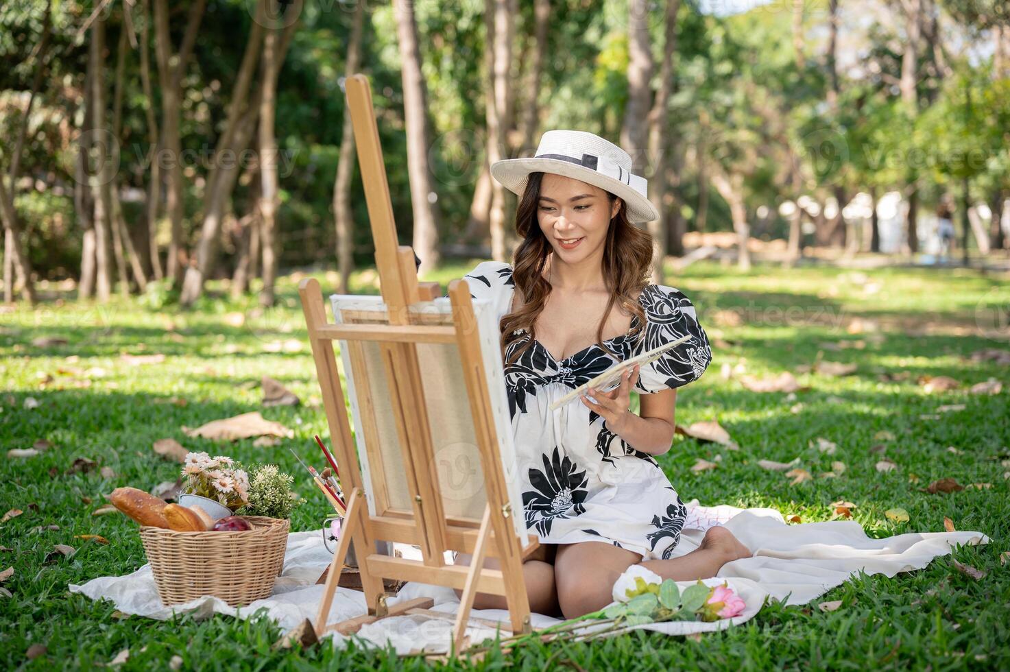 A charming Asian woman is painting a picture on a canvas easel while picnicking in a green park. photo