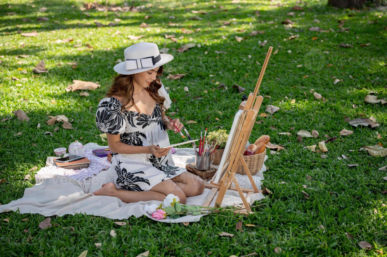 A beautiful Asian woman in a cute dress is painting on a canvas on an easel in a green park. photo