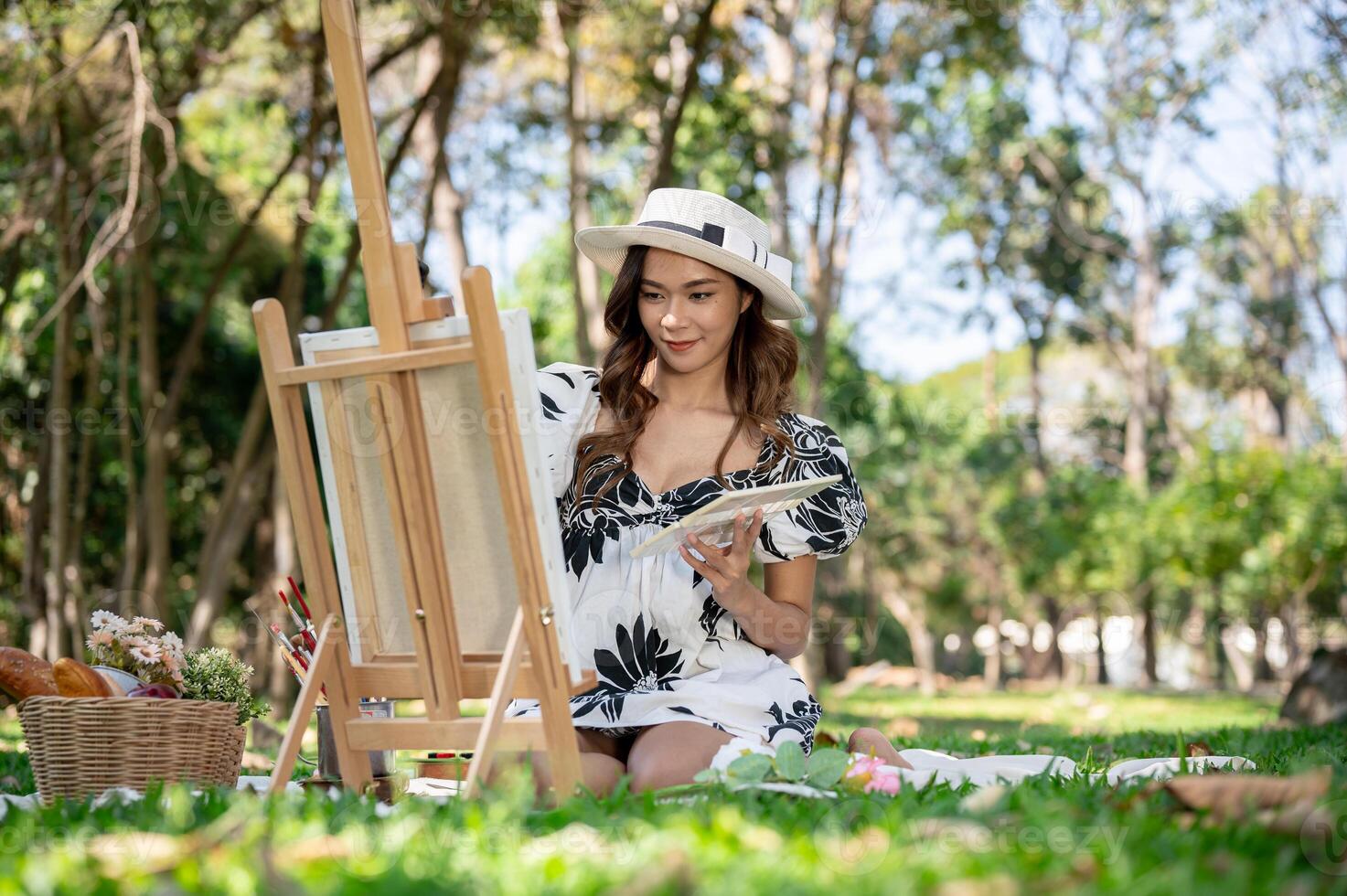 A charming Asian woman is painting a picture on a canvas easel while picnicking in a green park. photo