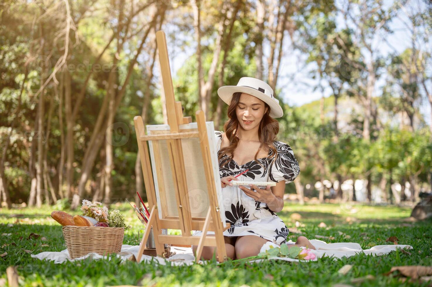 A charming Asian woman is painting a picture on a canvas easel while picnicking in a green park. photo