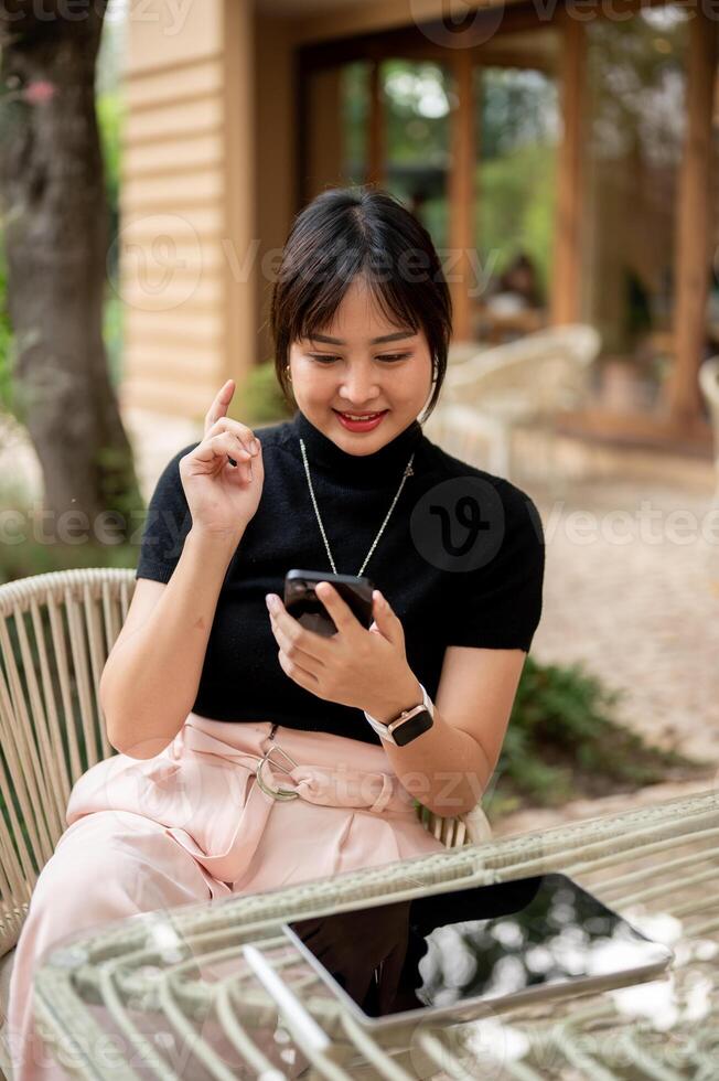 A cheerful, happy Asian woman sits at an outdoors table in the garden of a cafe using her smartphone photo