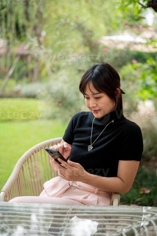 A beautiful Asian woman sits at a table in a green garden using her smartphone, checking messages. photo