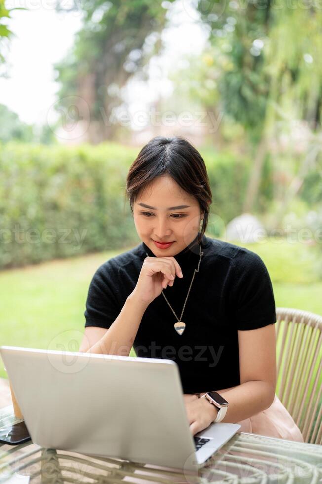 An attractive Asian woman sits at a table in a garden working, reading online document on her laptop photo