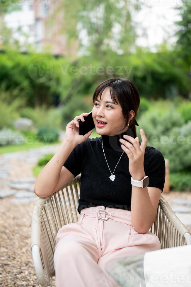 A positive Asian woman relaxing in a green garden, sitting at a table and talking on the phone. photo