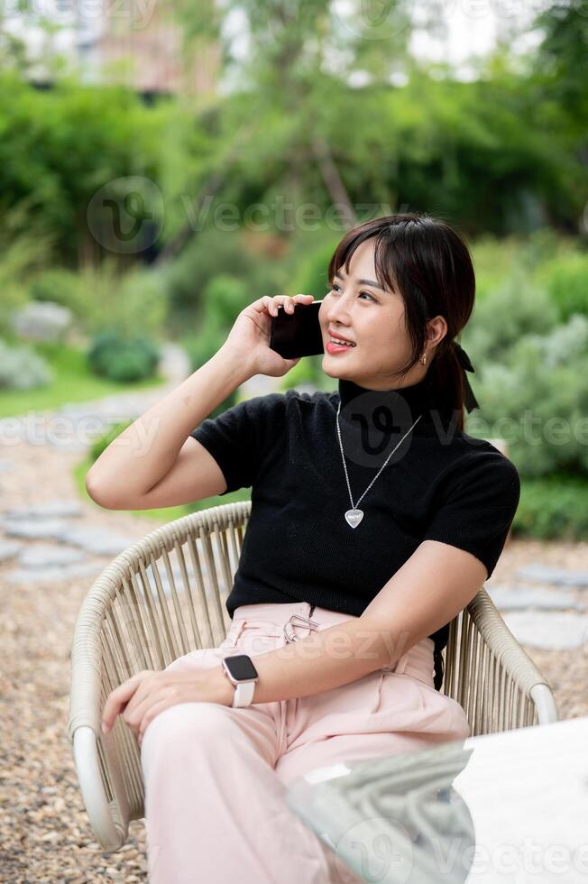 A positive Asian woman relaxing in a green garden, sitting at a table and talking on the phone. photo