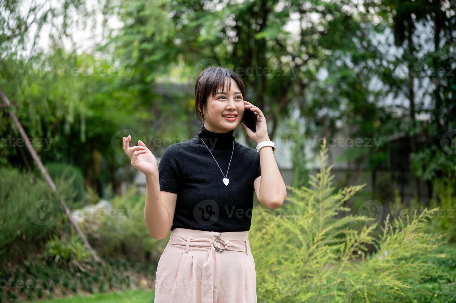 A positive, beautiful Asian woman talking on the phone while walking in a beautiful green garden. photo