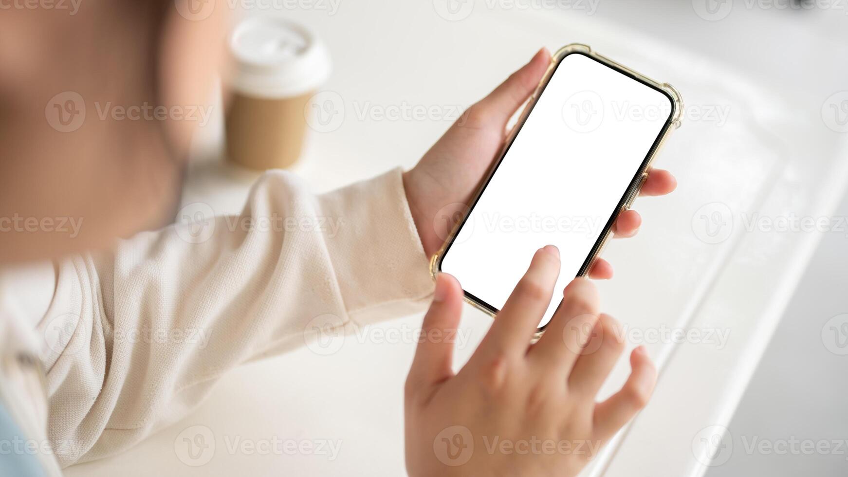 A woman in a white sweater holding a smartphone white-screen mockup over a white table indoors. photo