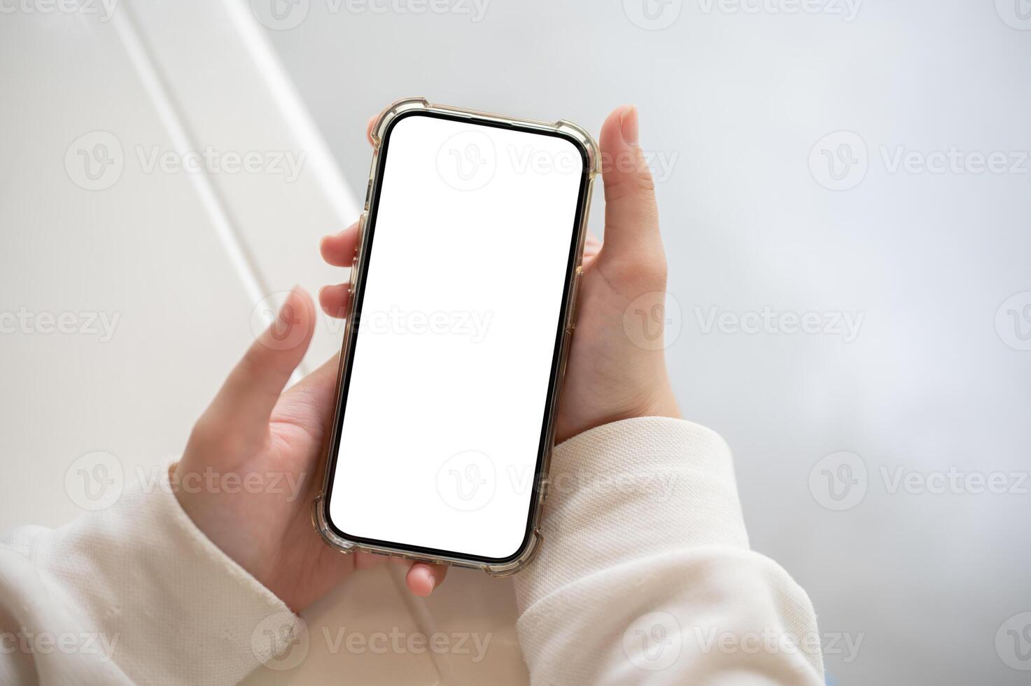 A woman in a white sweater holding a smartphone white-screen mockup over a white table indoors. photo