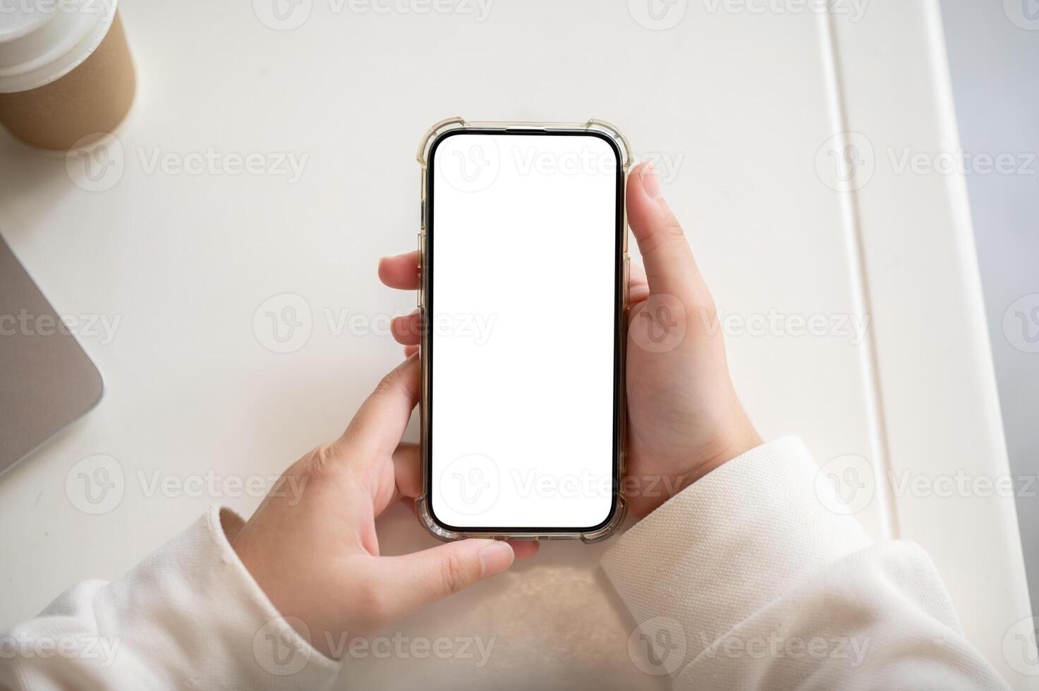 A woman in a white sweater holding a smartphone white-screen mockup over a white table indoors. photo