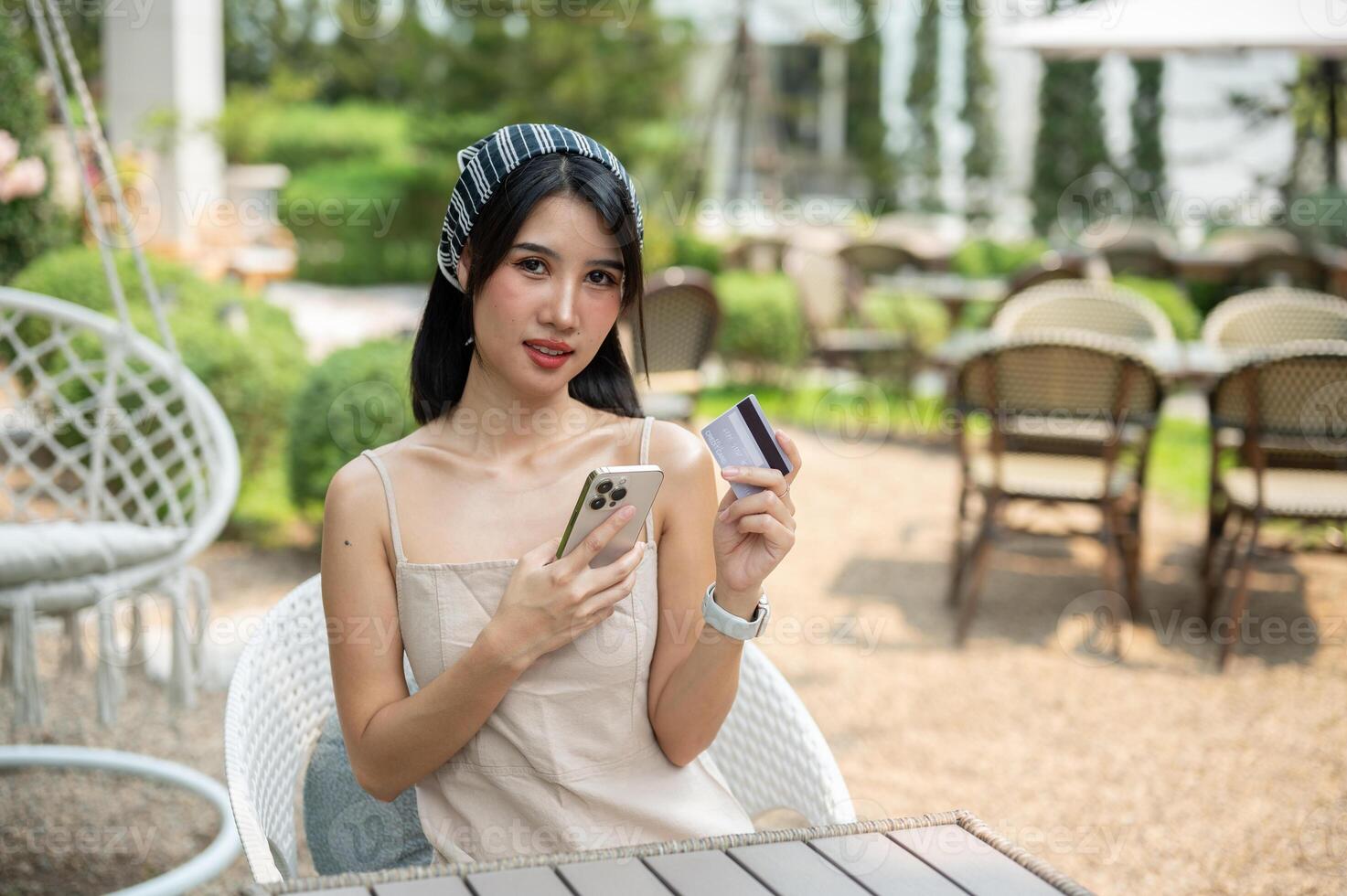 An attractive Asian woman sits at a table in a garden holding a credit card and smartphone. photo
