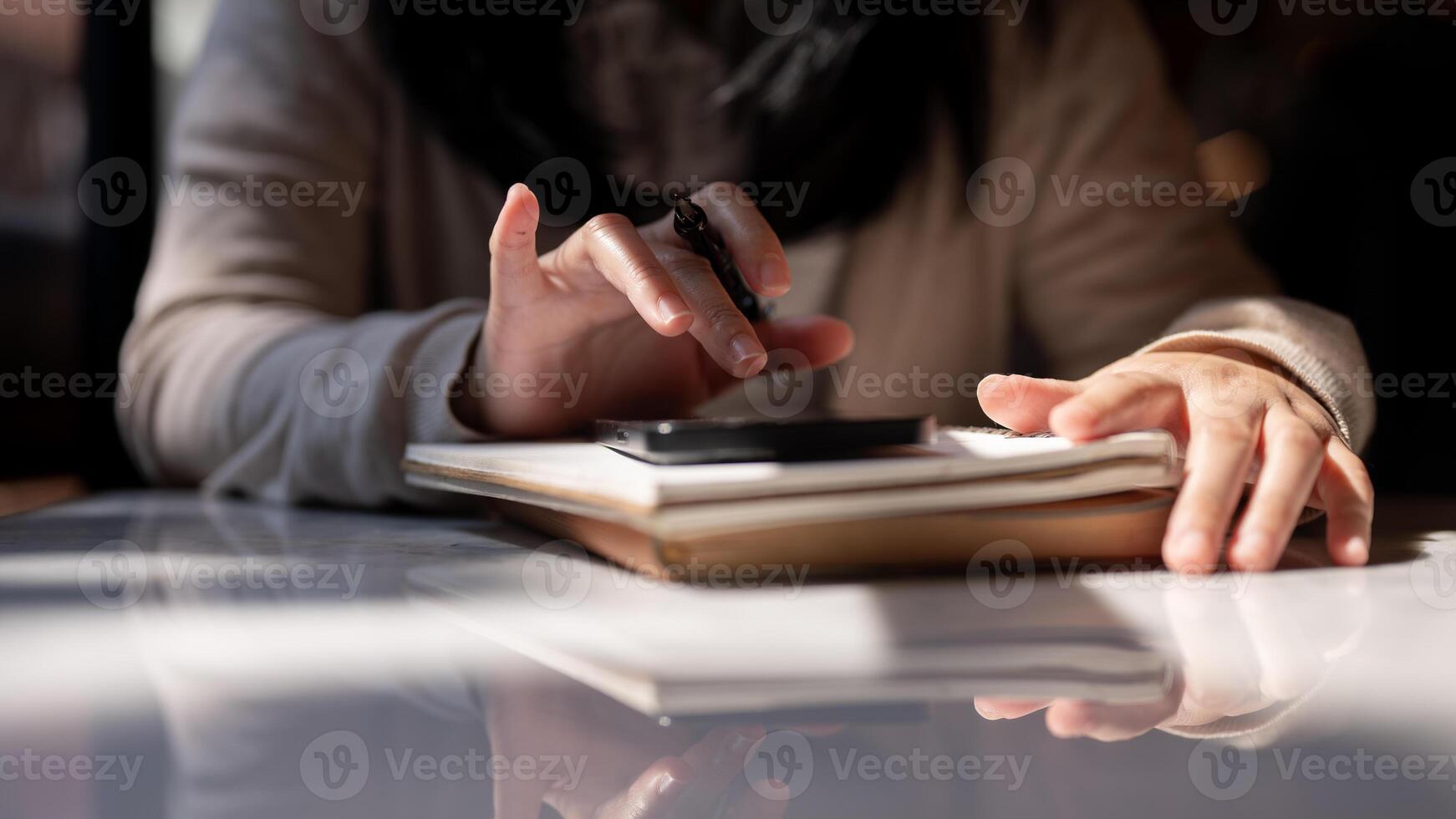 A cropped image of a woman holding a pen and using her smartphone at a table. photo
