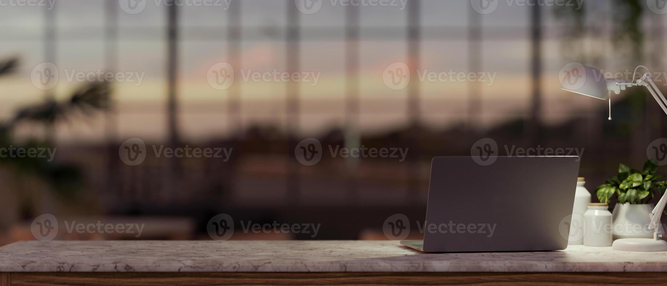 A laptop computer, a table lamp, and a potted plant are on a table in a modern dark office at sunset photo