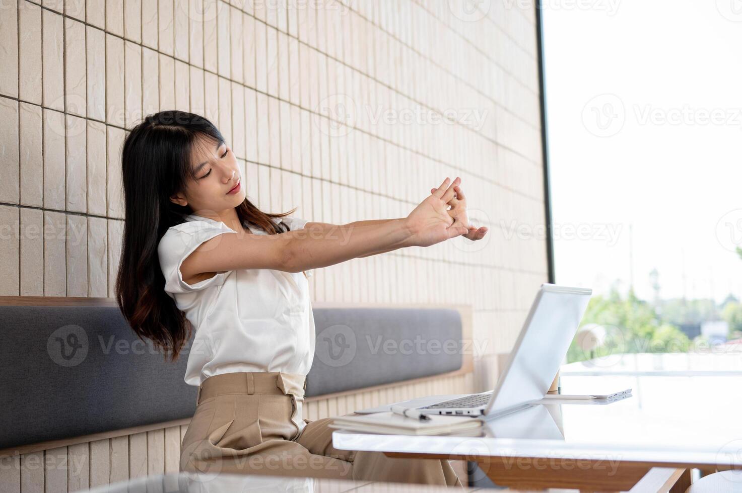 A beautiful Asian woman is feeling tired, stretching her hands while working remotely at a cafe. photo