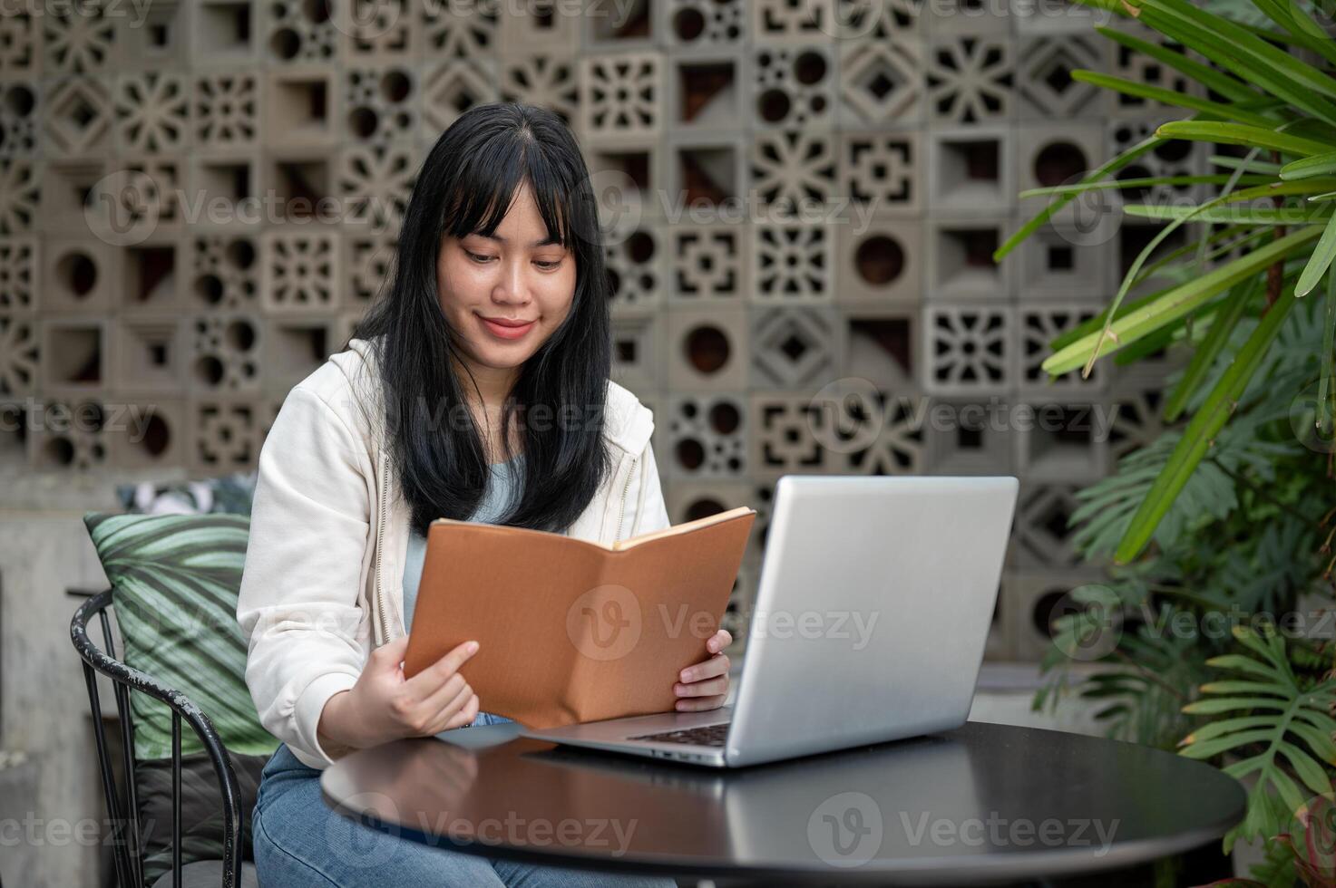 A young, positive Asian woman is reading a book at an outdoor table of a cafe, working remotely. photo