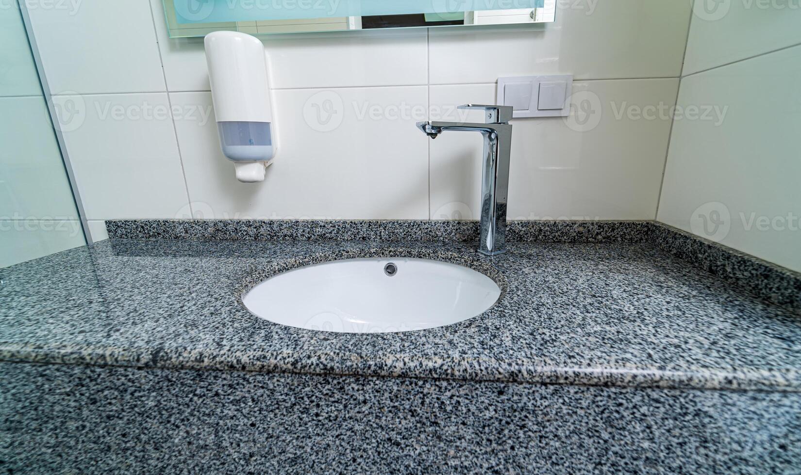 Public clinic bathroom with soap dispenser on tiled wall with mirror. Granite sink for washing hands and face. Closeup. photo