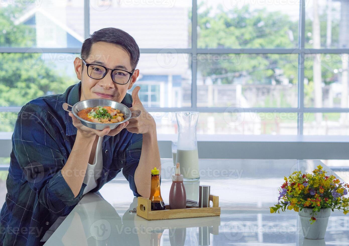 Young man wearing glasses holding the fried egg served on a pan with colorful toppings served on a pan while sitting on a chair. photo
