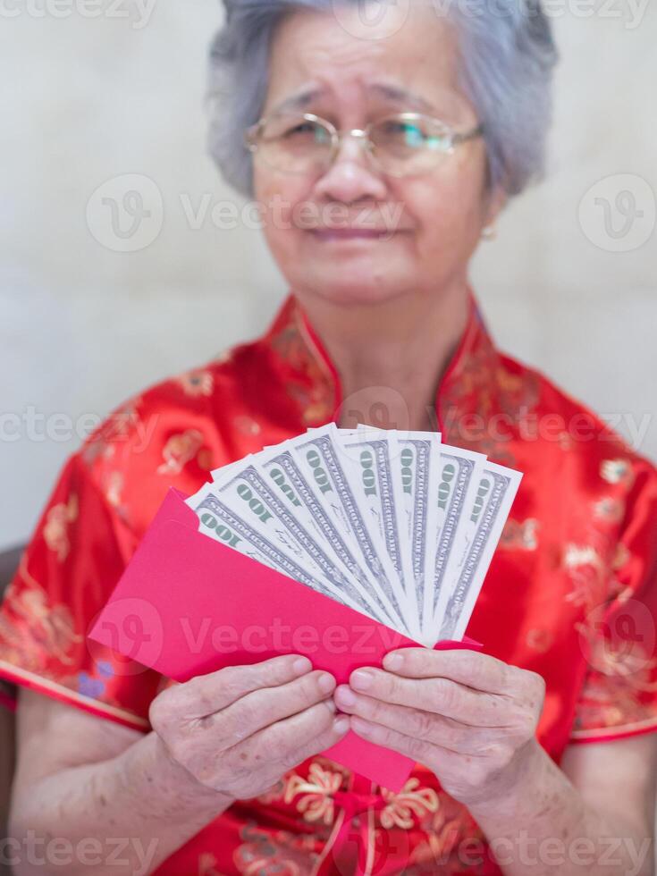 Senior woman wearing a traditional Cheongsam qipao dress holding US banknotes and looking at the camera while sitting on a chair. Concept of aged people and Chinese new year photo