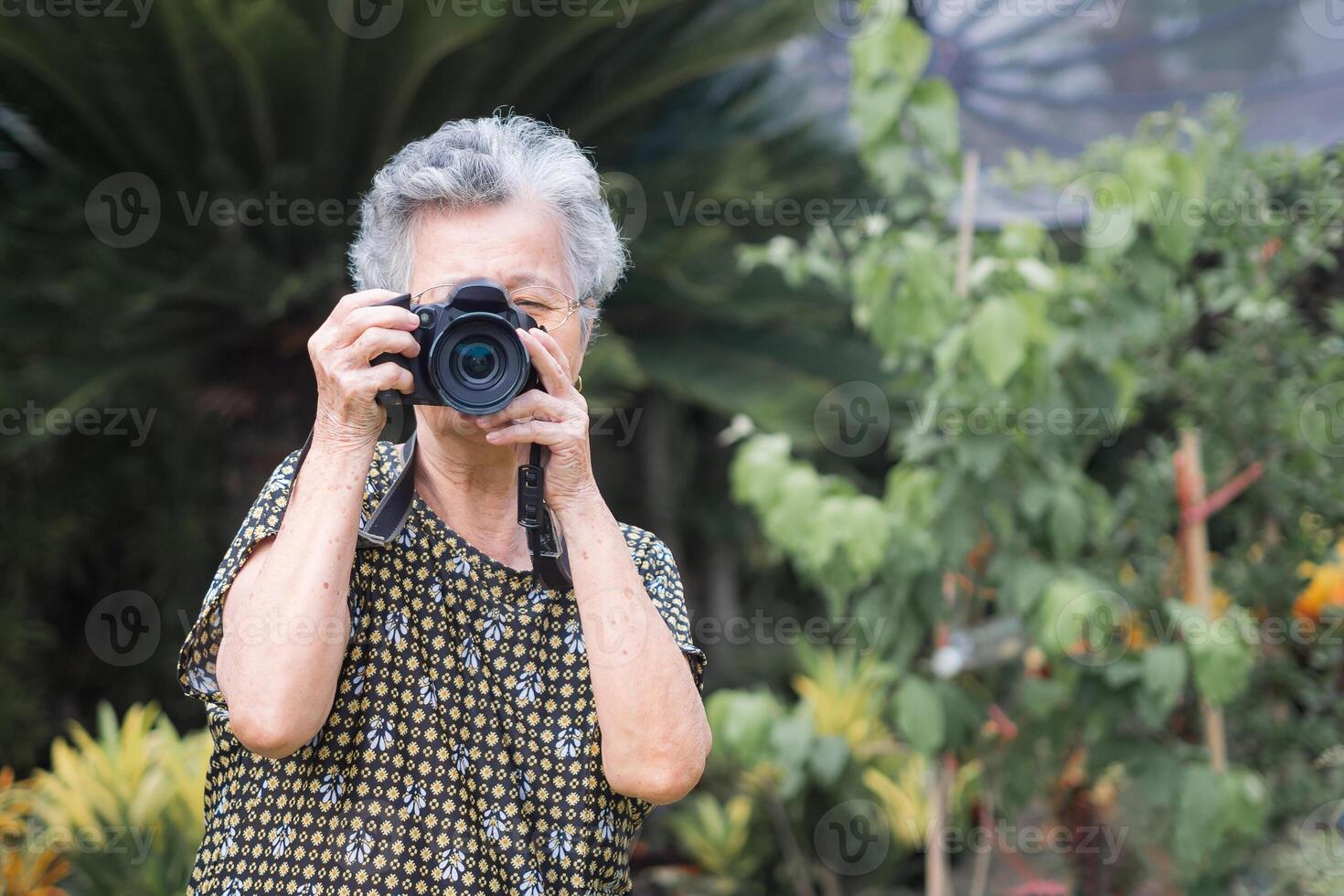 Senior woman with short gray hair shooting photos by a digital camera while standing outdoors. Space for text. Concept of aged people and photography