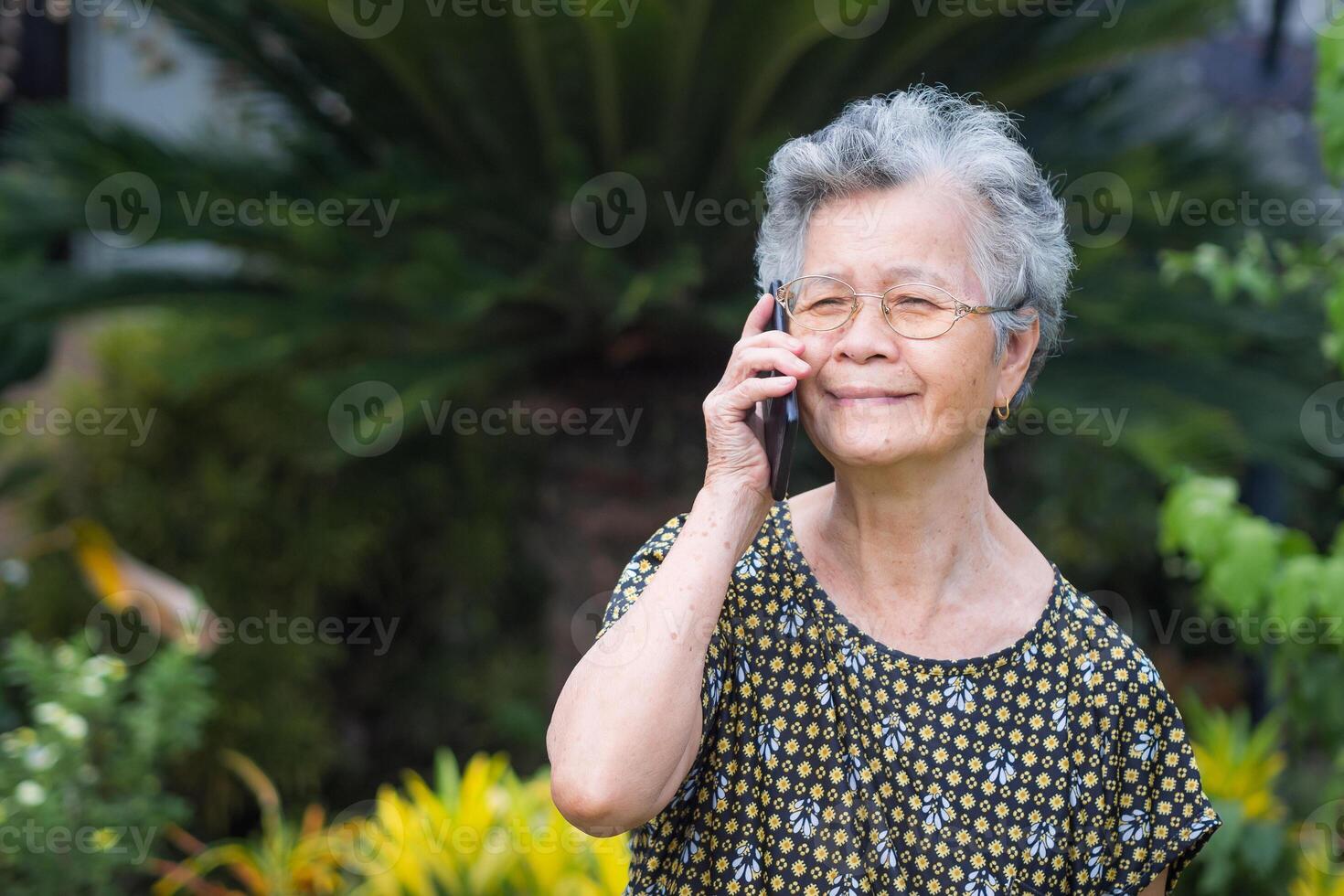 A portrait of a senior woman using a smartphone and looking away while standing in a garden. Space for text. Concept of aged people and technology photo