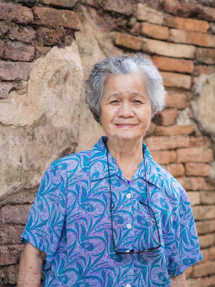 A portrait of an elderly Asian woman smiling and looking at the camera while standing with old brick wall background. Concept of aged people and relaxation photo