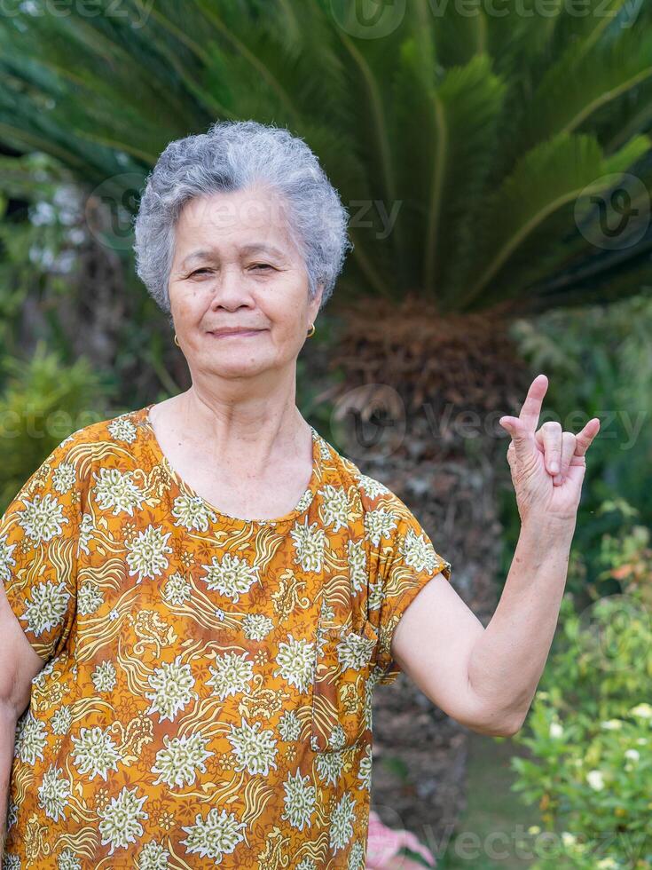 mayor mujer con corto gris pelo demostración dedos yo amor usted un símbolo, sonriente y mirando a el cámara mientras en pie en un jardín. concepto de Envejecido personas y cuidado de la salud foto