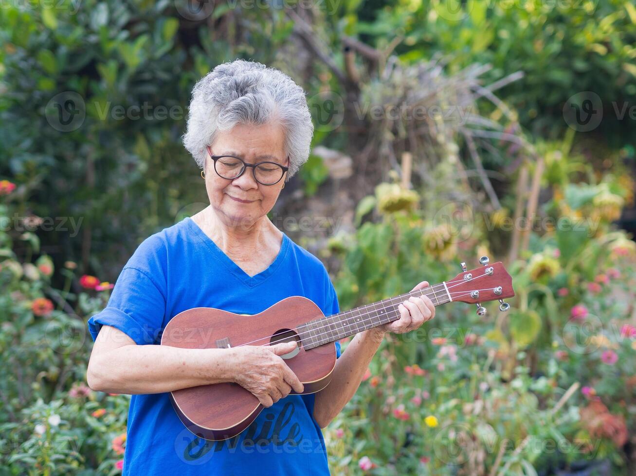 Portrait of elderly woman playing ukulele in her garden. Relaxing by singing and play small guitar happy and enjoy life after retired. Concept of old people and health care. Space for text photo