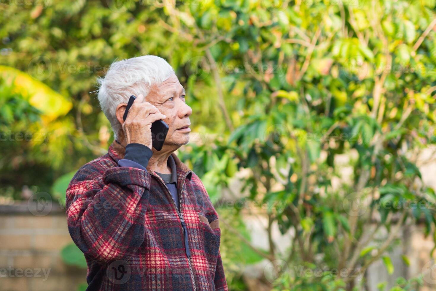 Elderly man using a smartphone and looking up while standing in a garden. Space for text. Concept of aged people and communication photo
