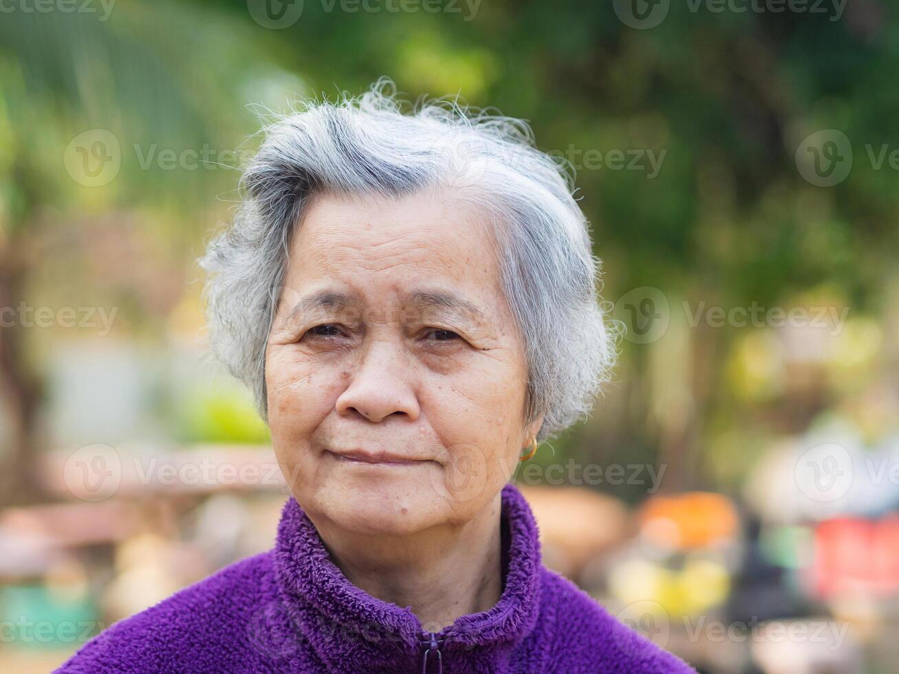 Portrait of elderly Asian woman with short gray hair and standing smiling and looking at the camera while standing in a garden. Space for text. Concept of aged people and relaxation photo