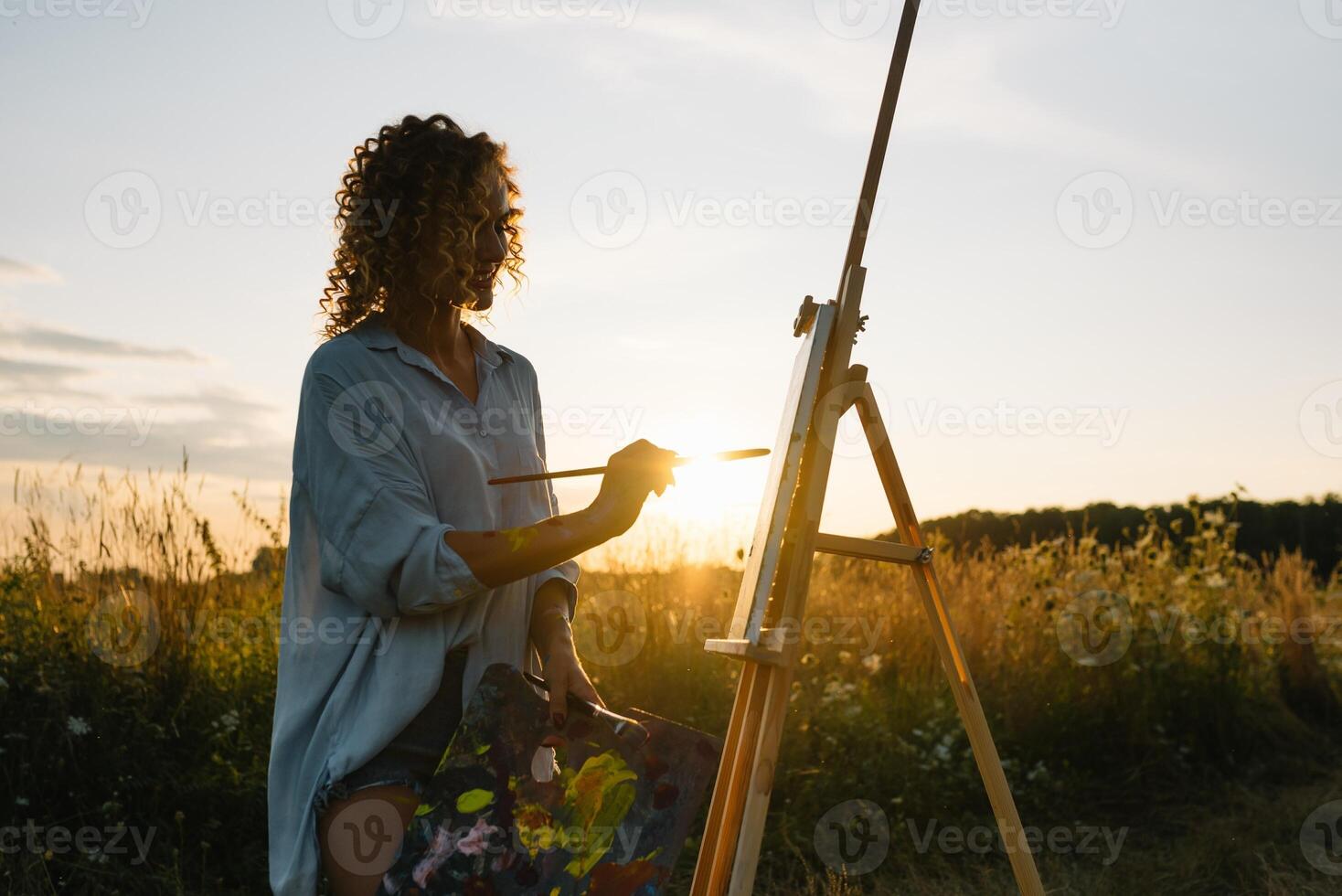Silhouette of a blonde girl artist. Lady paints a painting on the canvas with the help of paints. A wooden easel keeps the picture. Summer is a sunny day, sunset. photo