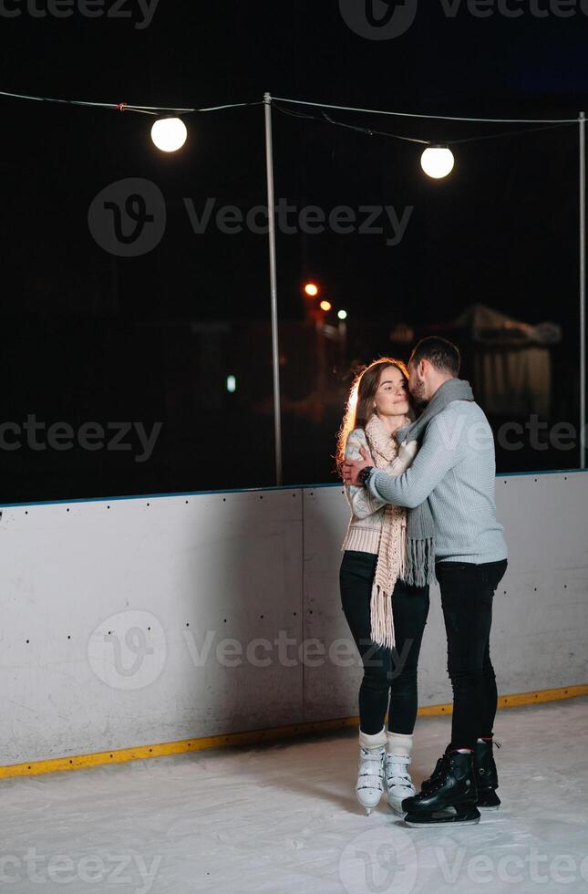 Theme ice skating rink and loving couple. meeting young, stylish people ride by hand in crowd on city skating rink lit by light bulbs and lights. Ice skating in winter for Christmas on ice arena. photo