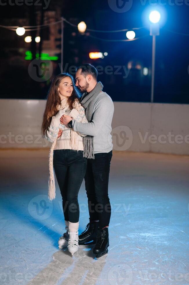 Couple on the city rink in a winter evening. Guy helping nice girl to skate on the ice in the dark night and twinkles lighting above them photo