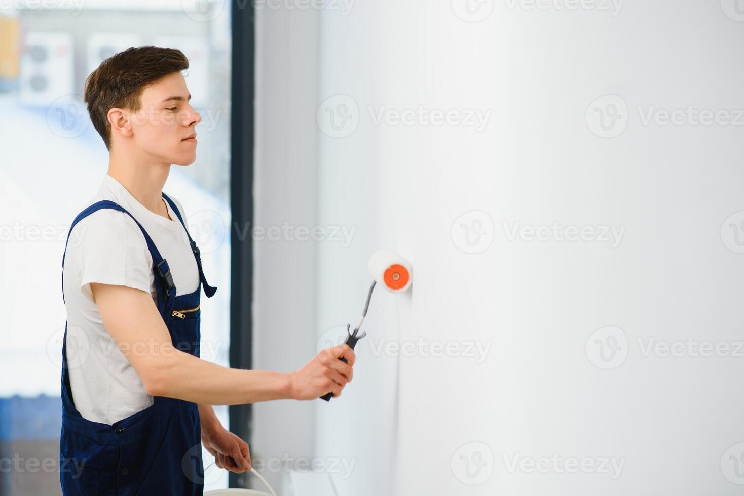young man in blue overalls painting wall into white color with a roller photo