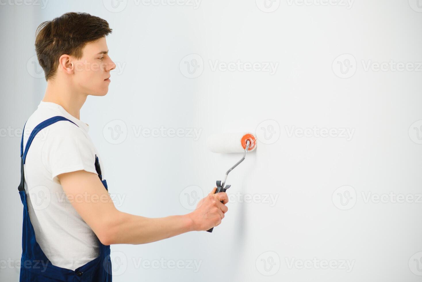 young man in blue overalls painting wall into white color with a roller photo