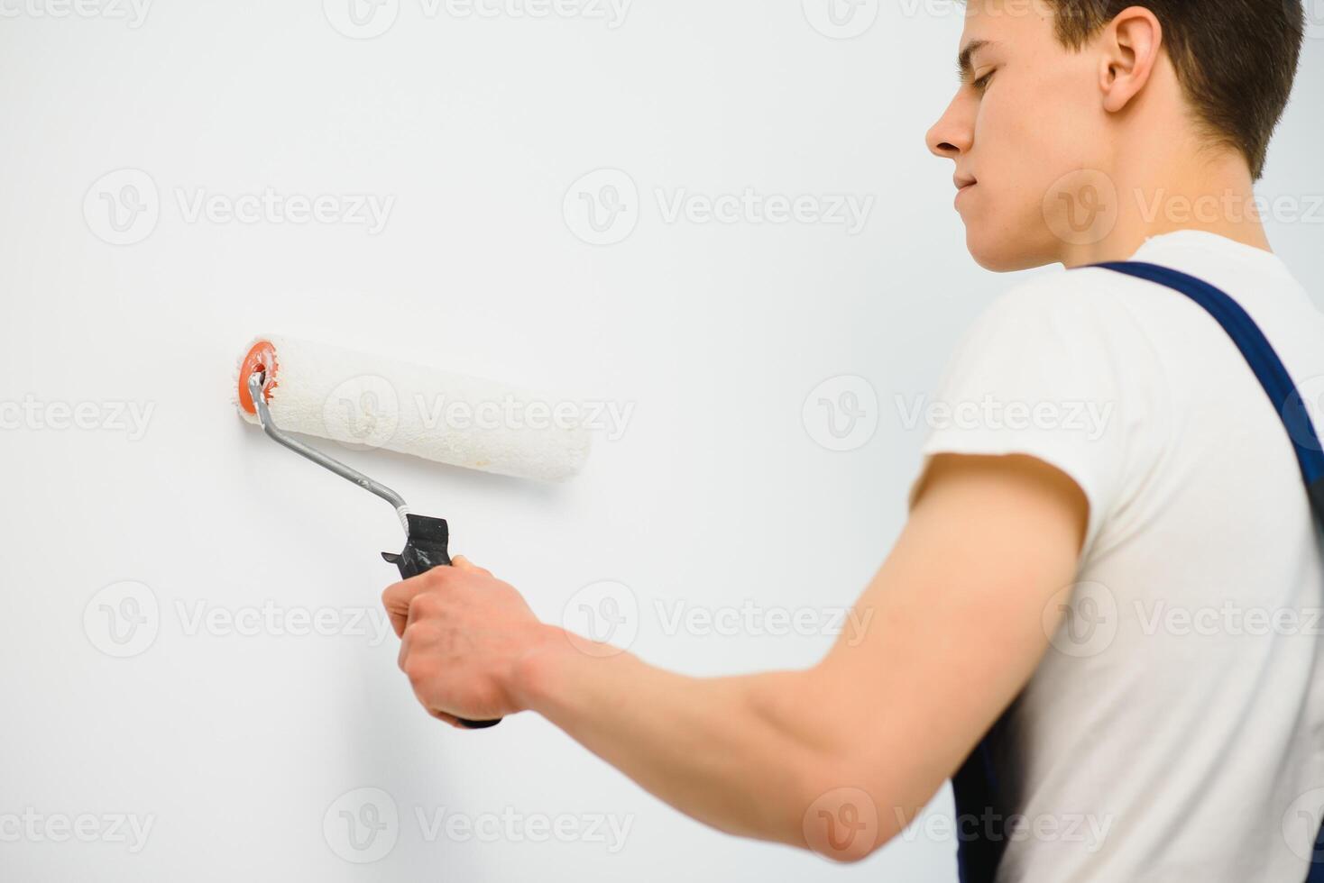 young man in blue overalls painting wall into white color with a roller photo