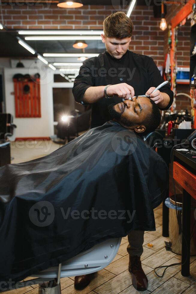 Side view of serious man with stylish modern haircut looking forward in barber shop. Hand of barber keeping straight razor and cutting trendy stripes on head of client. photo