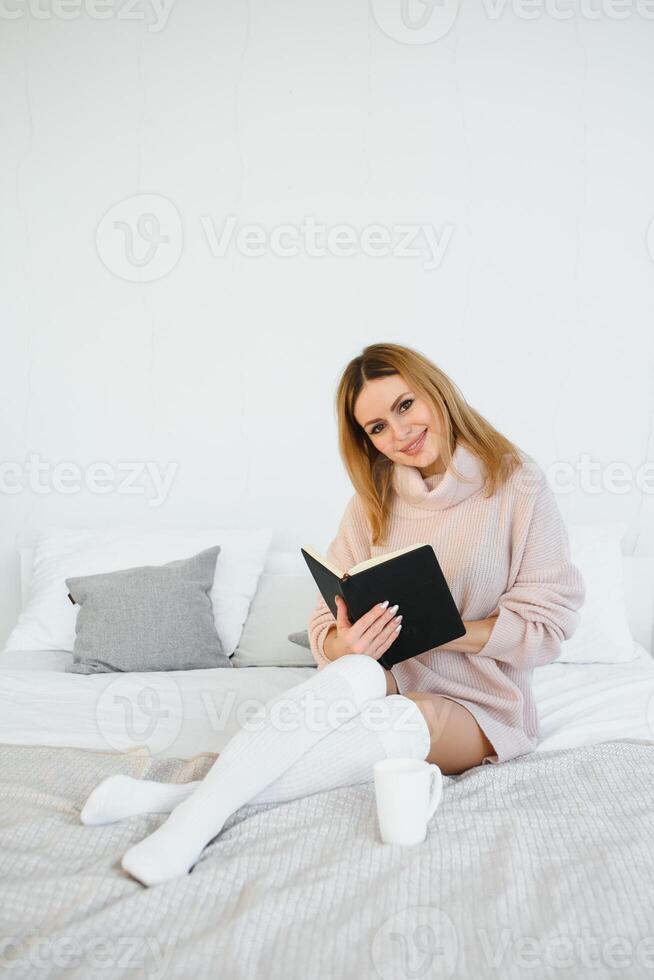 A woman reading a book and smiling as she sits in bed. The alarm clock on the desk beside her. photo