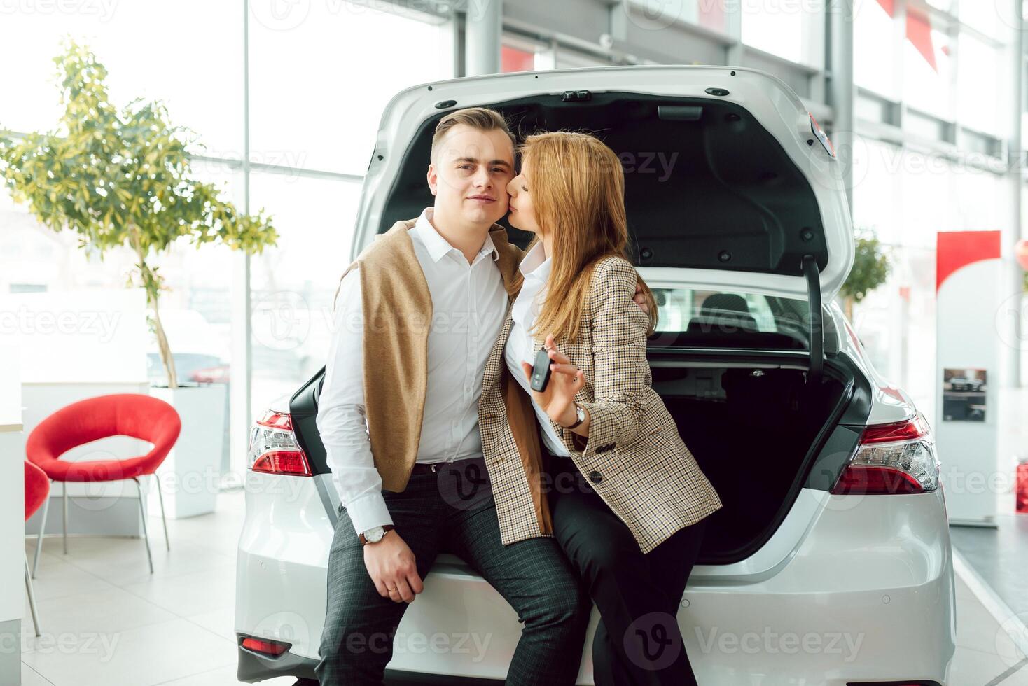 Visiting car dealership. Beautiful couple is holding a key of their new car and smiling, girl is kissing her husband in cheek. photo