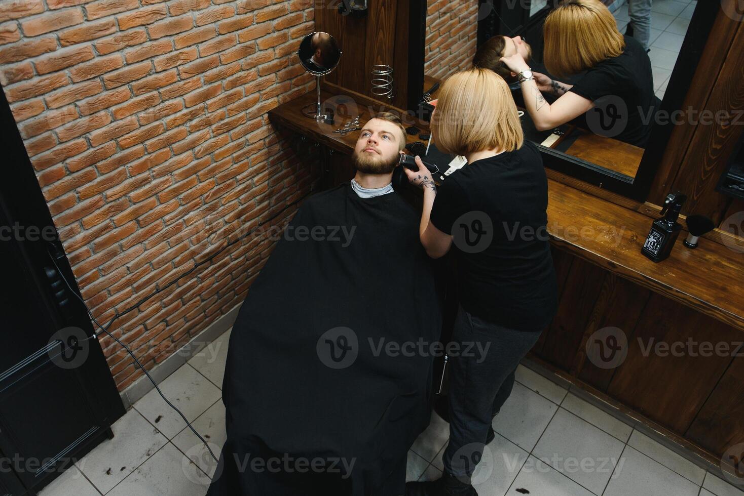 Skilled female barber cutting a beard of brunette adult guy in professional retro barbershop. photo