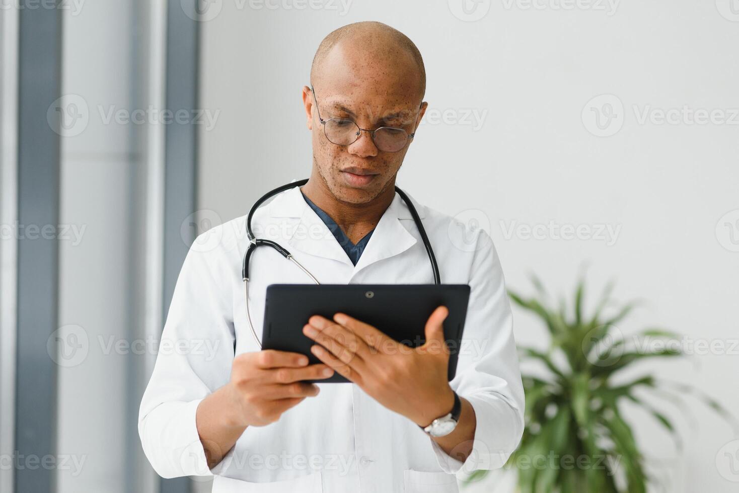 Mature african doctor using digital tablet in corridor . Portrait of confident male doctor using tablet computer in clinic with copy space. Successful smiling doctor in labcoat wearing stethoscope photo