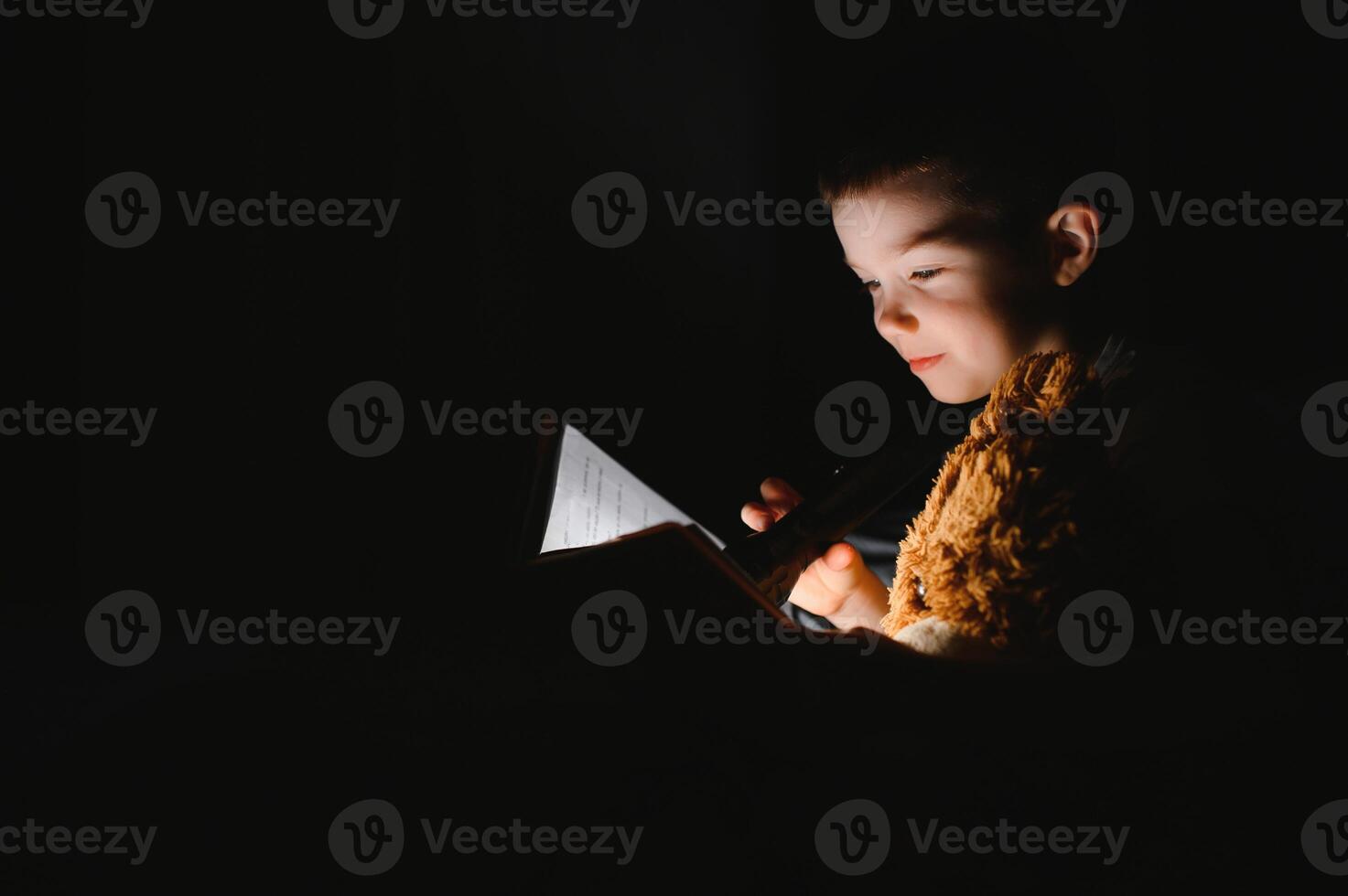 Boy with flashlight reading book under blanket at home. photo