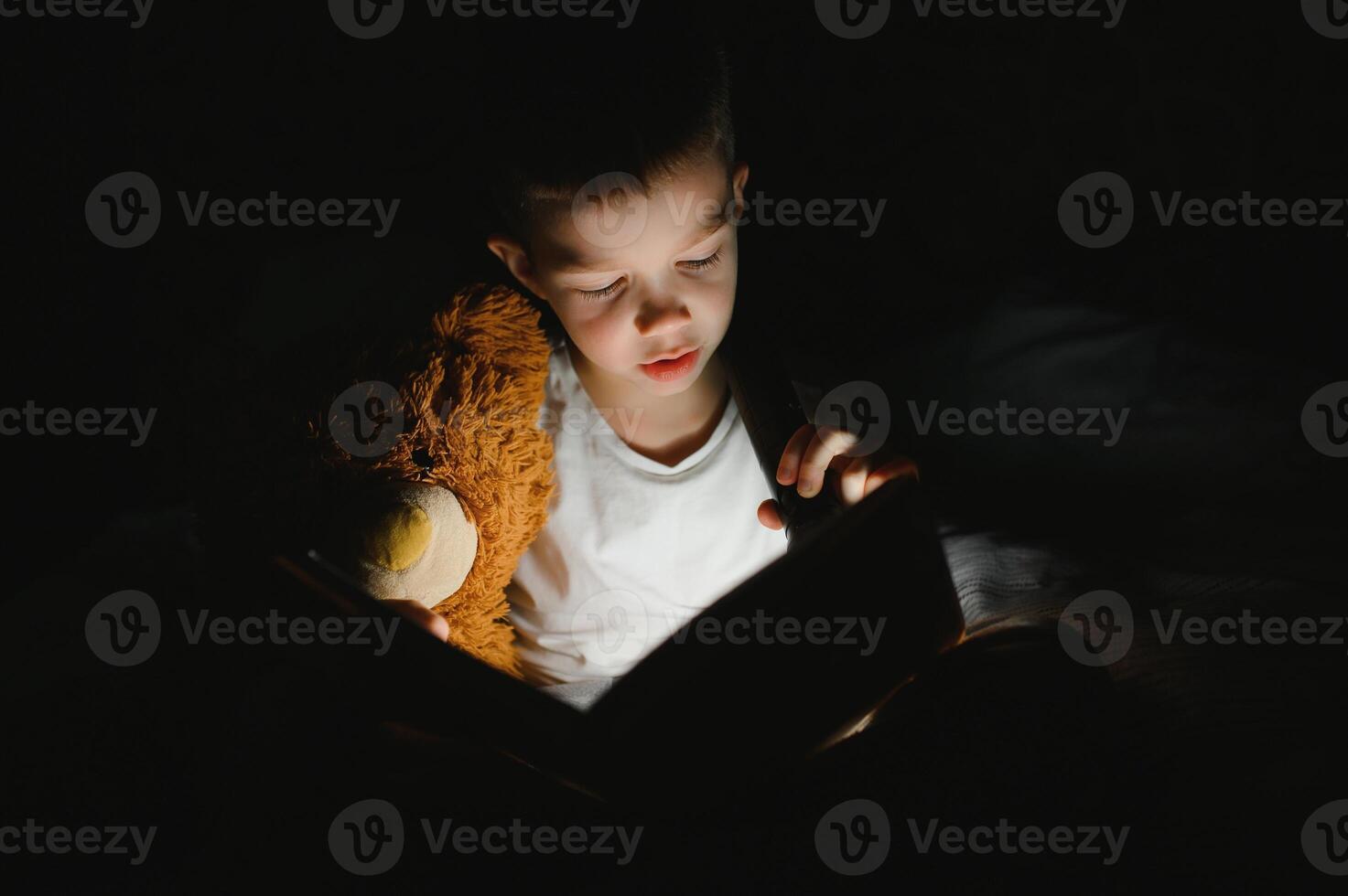 Boy with flashlight reading book under blanket at home. photo