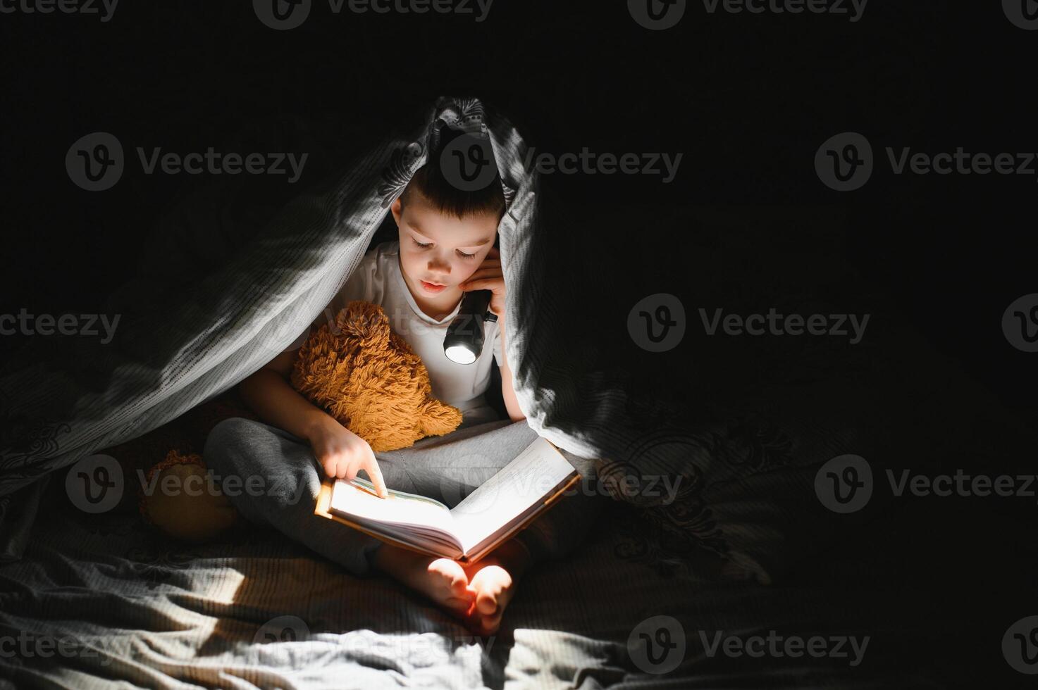 Boy with flashlight reading book under blanket at home. photo