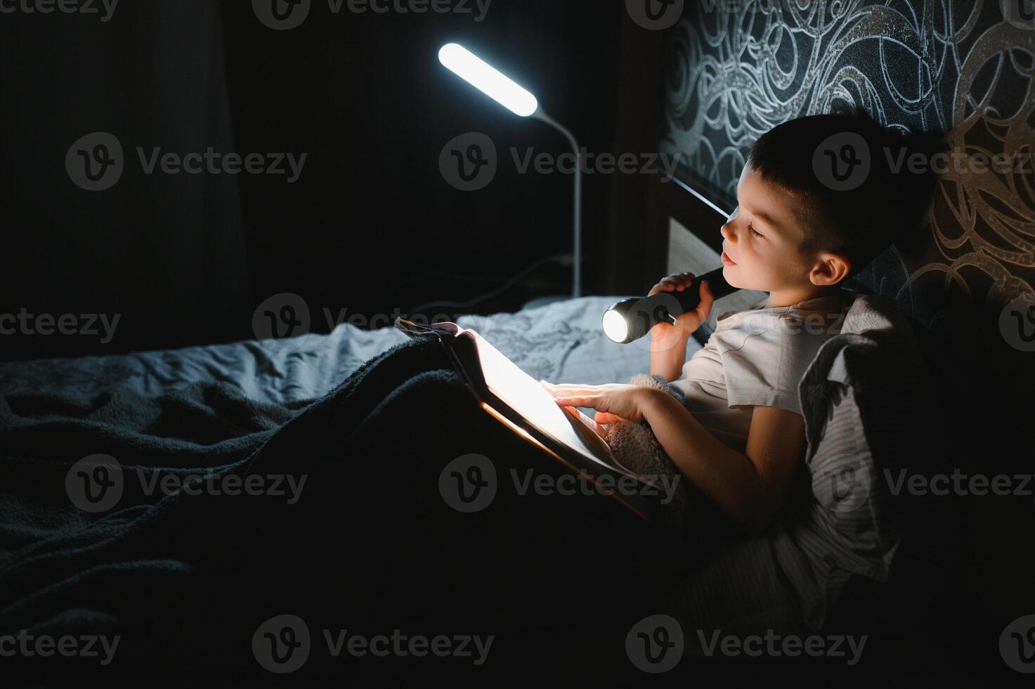Child reading book in bed. Kids read at night. Little boy with fairy tale books in bedroom . Education for young children. Bedtime story in the evening. Cute kid under blanket in dark room with lamp photo