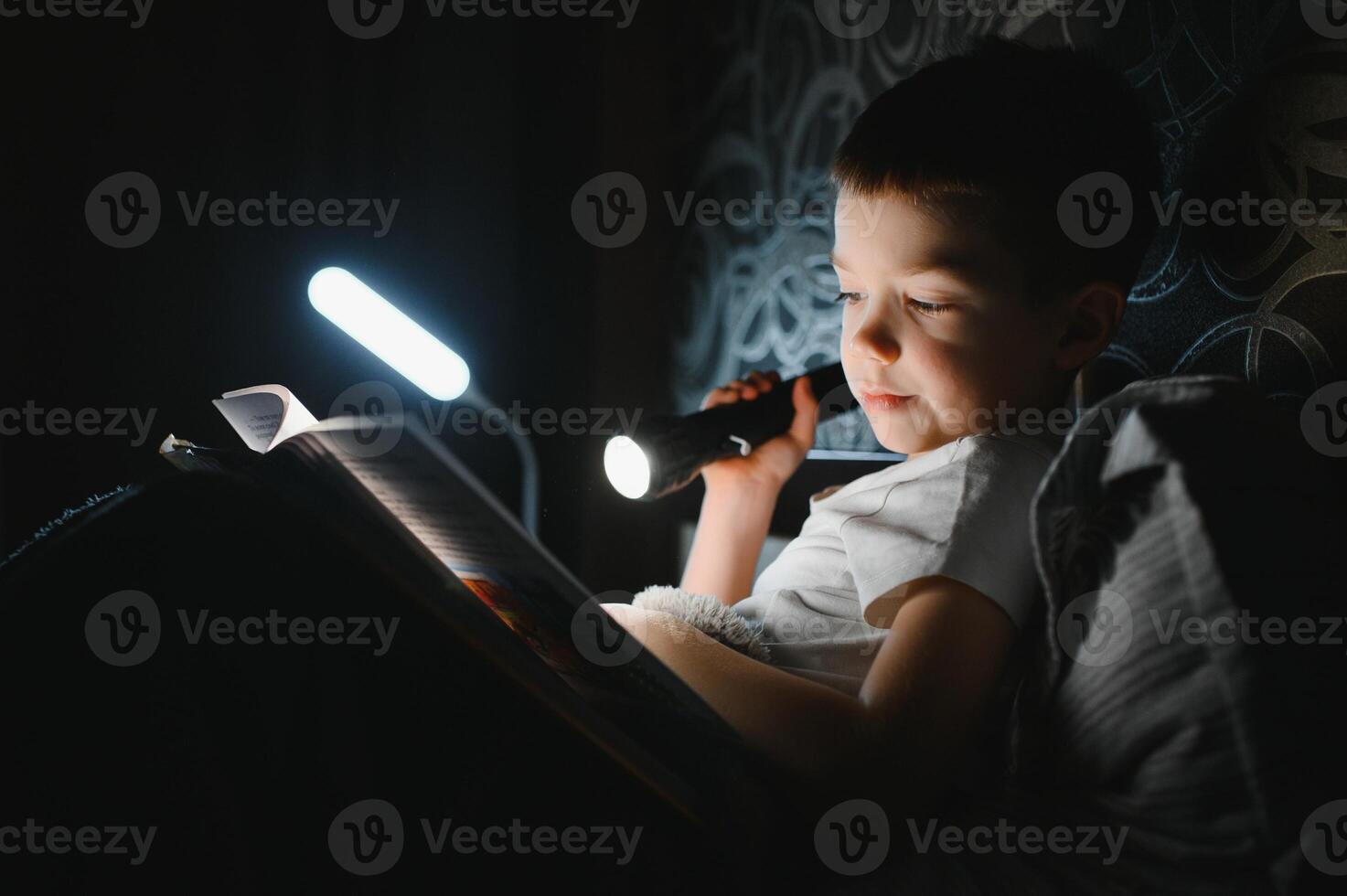 Boy with flashlight reading book under blanket at home. photo
